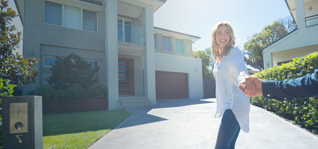 Frau zieht einen Mann, der nur halb auf dem Bild zu sehen ist, an der Hand in Richtung eines Einfamilienhauses – Symbolbild Grundbesitzerhaftpflicht