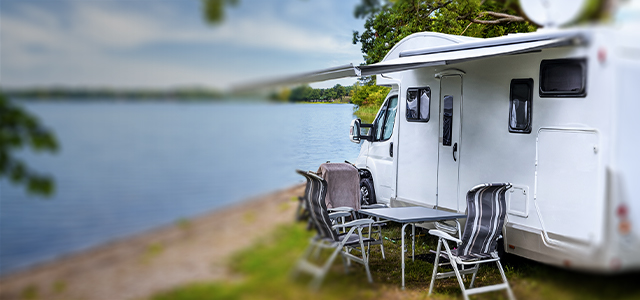 Campingmobil mit Tisch und Stühlen davor steht am Ufer eines Sees – Symbolbild für eine Kfzversicherung für Camper und Wohnwagen