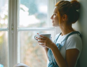 Frau sitzt gemütlich mit einer Tasse Tee in der Hand vor einem Fenster und schaut hinaus – Symbolbild für eine Glasversicherung