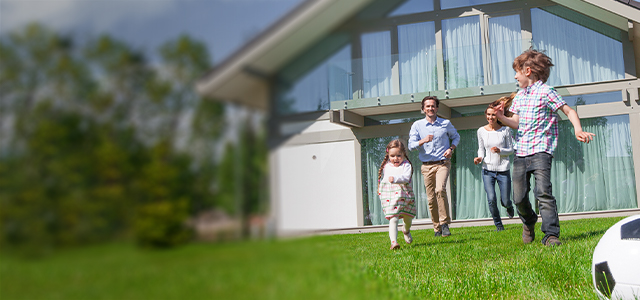 Familie spielt auf dem Rasen vor einem modernen Einfamilienhaus bei Sonnenschein – Symbolbild für eine Wohngebäudeversicherung