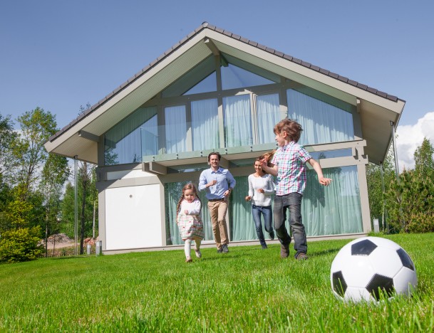 Familie spielt auf dem Rasen vor einem modernen Einfamilienhaus bei Sonnenschein – Symbolbild für eine Wohngebäudeversicherung