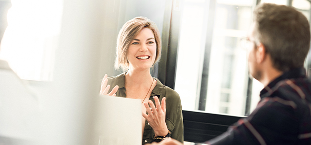 Eine Frau vor einem Laptop spricht gestikulierend mit einem Mann von dem man nur die Rückansicht sieht