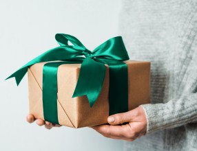 Ein Geschenkpaket mit einer grünen Schleife, wird von einer Person gehalten von der man nur die Hände sieht – Symbolbild für vermögenswirksame Leistungen